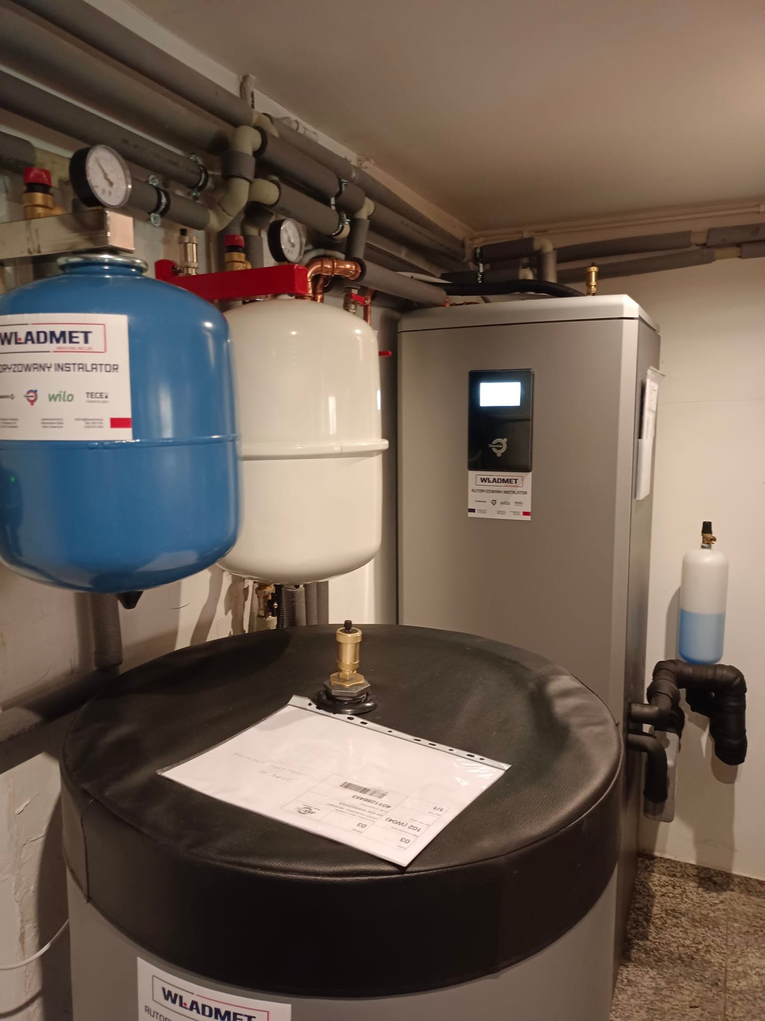 Instalacja grzewcza w kotłowni z dwoma zbiornikami wyrównawczymi (niebieski i biały) oraz szarym zasobnikiem ciepłej wody, widoczne rury, manometry i etykieta firmy Władmet na urządzeniach.