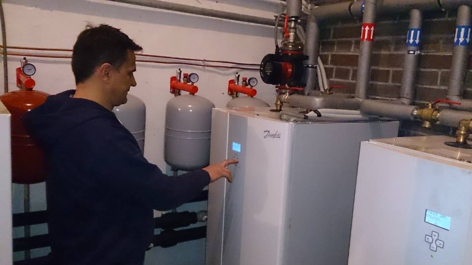 Mężczyzna w bluzie z kapturem obsługuje panel sterowania białej instalacji grzewczej Danfoss w kotłowni z rurami i zbiornikami.