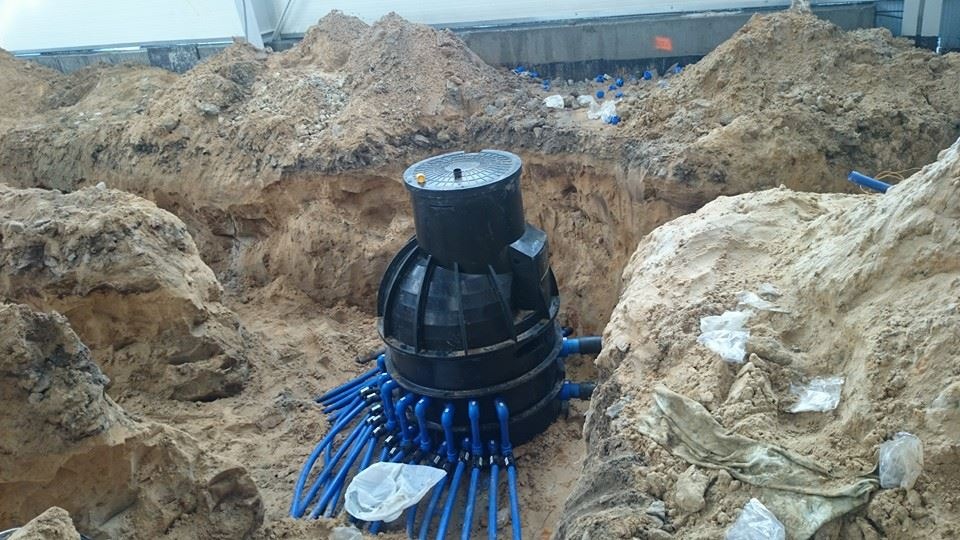 Czarna, segmentowa konstrukcja z podłączonymi niebieskimi rurami, umieszczona w wykopie ziemnym, widok z góry na krawędź wykopu i fragment hali w tle.