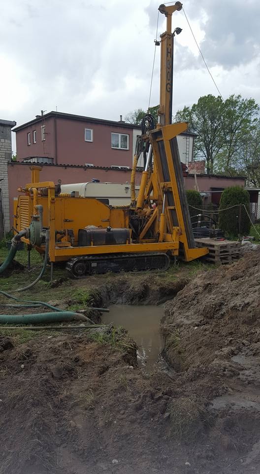 Żółta wiertnica na gąsienicach stoi na placu budowy obok wykopu wypełnionego wodą, w tle budynek mieszkalny i drzewa.