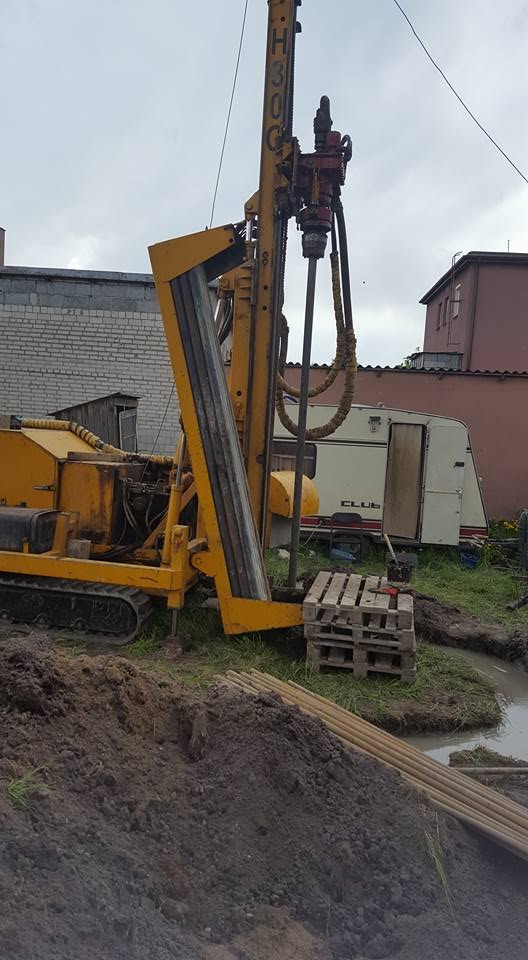Żółta wiertnica H30C podczas prac ziemnych, widoczne pale i teren budowy z przyczepą kempingową w tle.