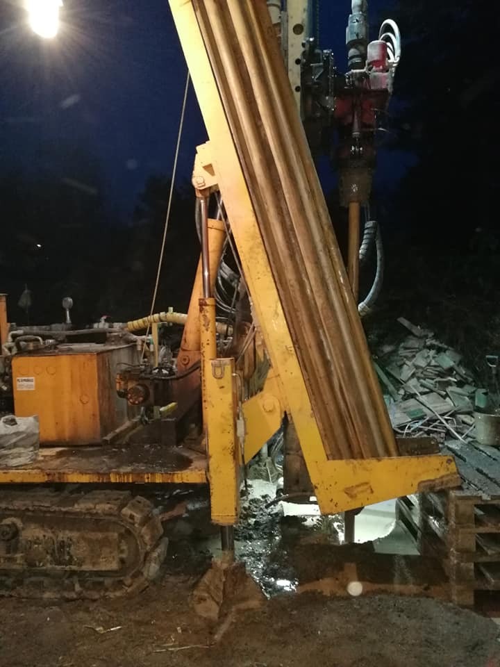 Żółta wiertnica na gąsienicach, w trakcie wiercenia otworu w ziemi, widoczne błoto i oświetlenie nocne.