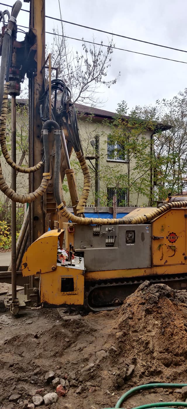 Żółta wiertnica na gąsienicach na placu budowy z starym budynkiem w tle.