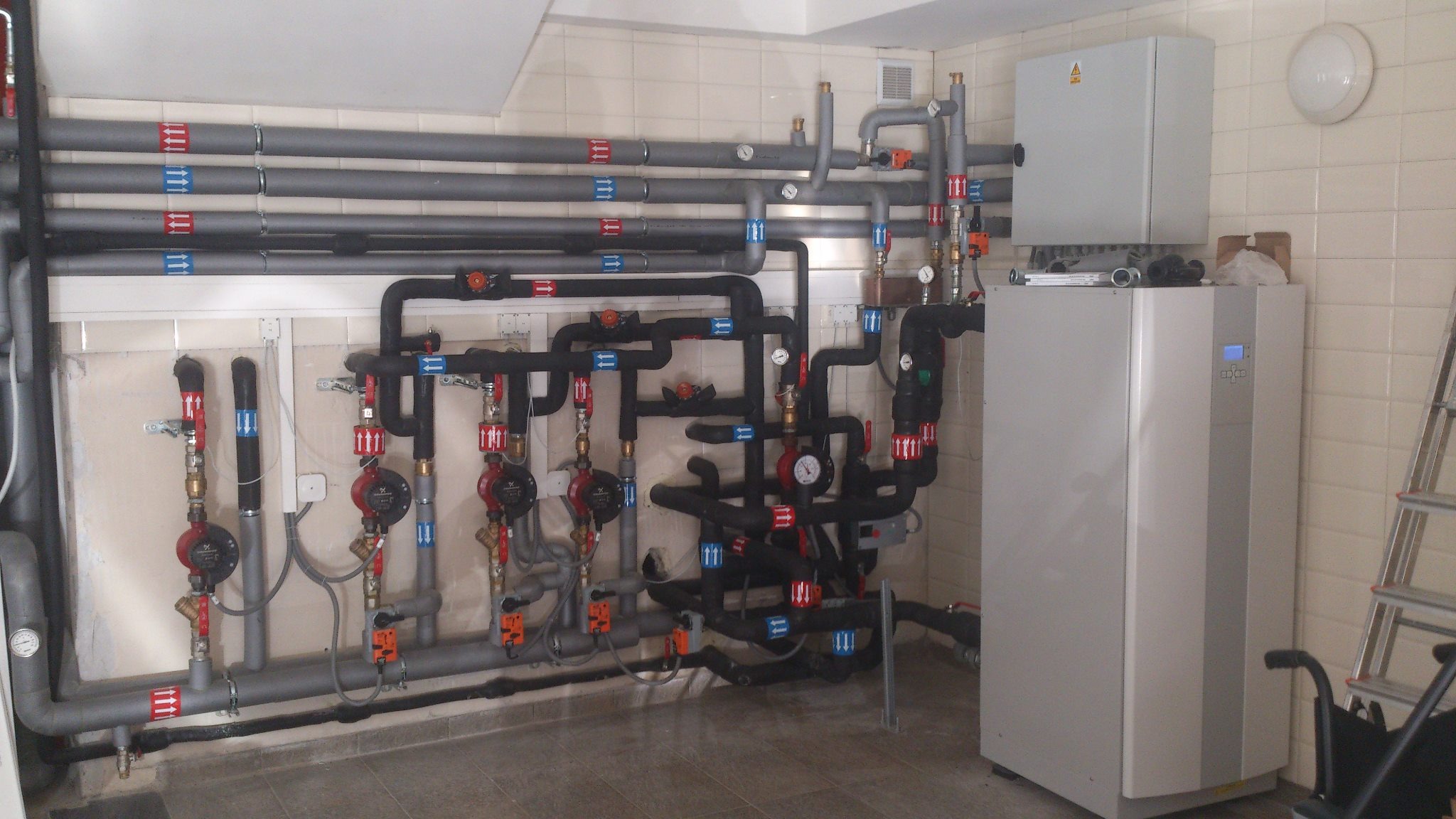 Instalacja grzewcza z licznymi rurami w kolorze szarym i czarnym, z czerwonymi i niebieskimi oznaczeniami kierunku przepływu, oraz szara skrzynka i urządzenie z wyświetlaczem na tle beżowych płytek...