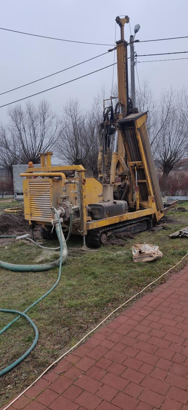Żółta wiertnica na gąsienicach z przewodami, w trakcie wiercenia otworów pod instalację pompy ciepła na trawniku obok czerwonej kostki brukowej.