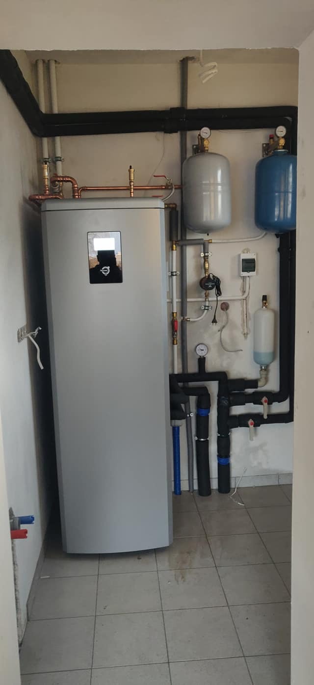 Szara jednostka wewnętrzna z wyświetlaczem, instalacja rur miedzianych i czarnych rur izolowanych, dwa zbiorniki ciśnieniowe (szary i niebieski) oraz manometr w pomieszczeniu z kafelkami na podłodze.