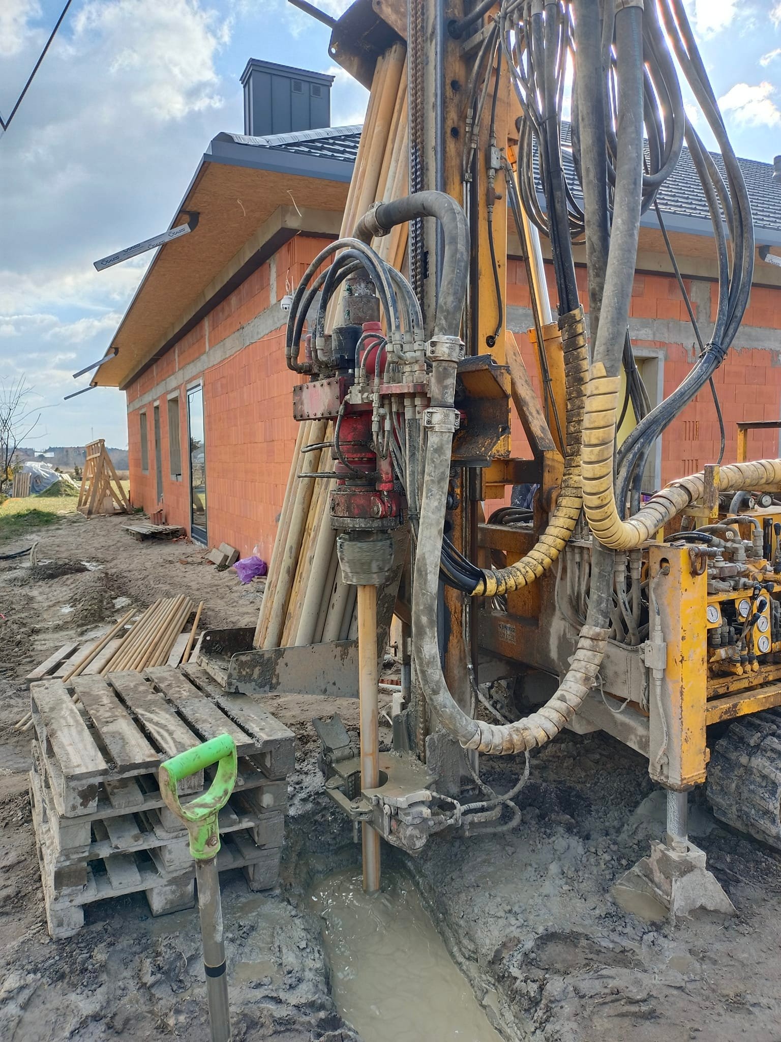 Wiertnica geologiczna w trakcie pracy przy budowie domu z czerwonej cegły, widoczna zielona łopata oparta o paletę