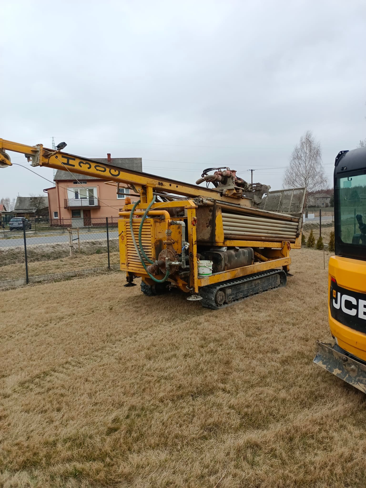 Żółta wiertnica H300 na gąsienicach, na trawiastym terenie, obok koparki JCB, z widocznym budynkiem mieszkalnym w tle.