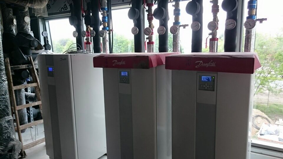 Trzy białe urządzenia Danfoss z czerwonymi górnymi panelami, prawdopodobnie jednostki grzewcze, stojące w pomieszczeniu technicznym z rurami i oknem z widokiem na zewnątrz.