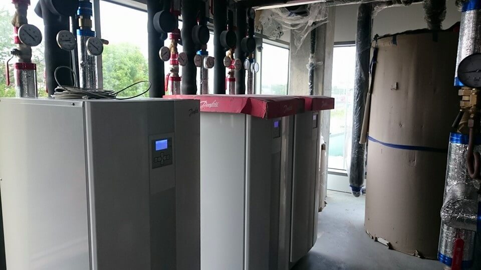 Rząd białych jednostek z manometrami i rurami, prawdopodobnie w kotłowni, z widocznym logo Danfoss na górze jednej z jednostek.