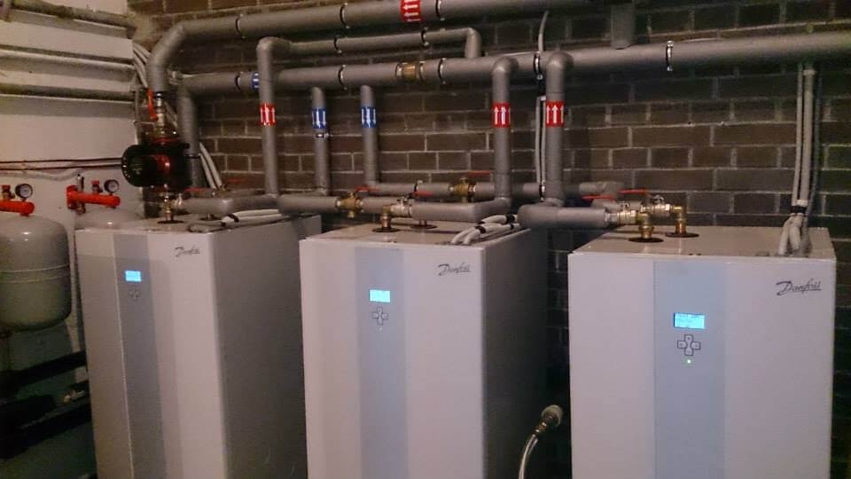 Trzy białe jednostki grzewcze Danfoss z wyświetlaczami, połączone rurami w instalacji w pomieszczeniu z ceglaną ścianą.