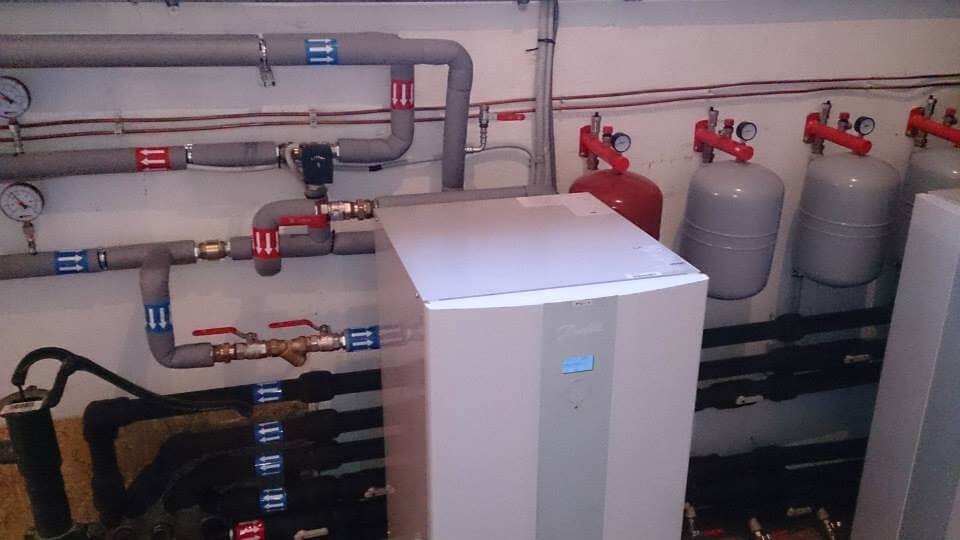 Instalacja grzewcza z szarymi rurami oznaczonymi strzałkami, zbiornikami wyrównawczymi i jednostką Danfoss z niebieskim wyświetlaczem.