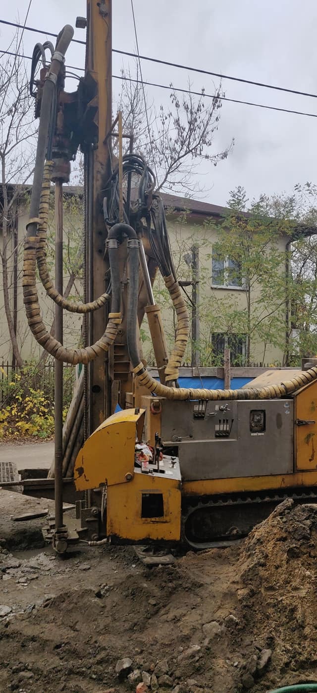 Żółta wiertnica do pomp ciepła na gąsienicach, z widocznymi przewodami i elementami sterującymi, w trakcie wiercenia w ziemi, z budynkiem mieszkalnym w tle.