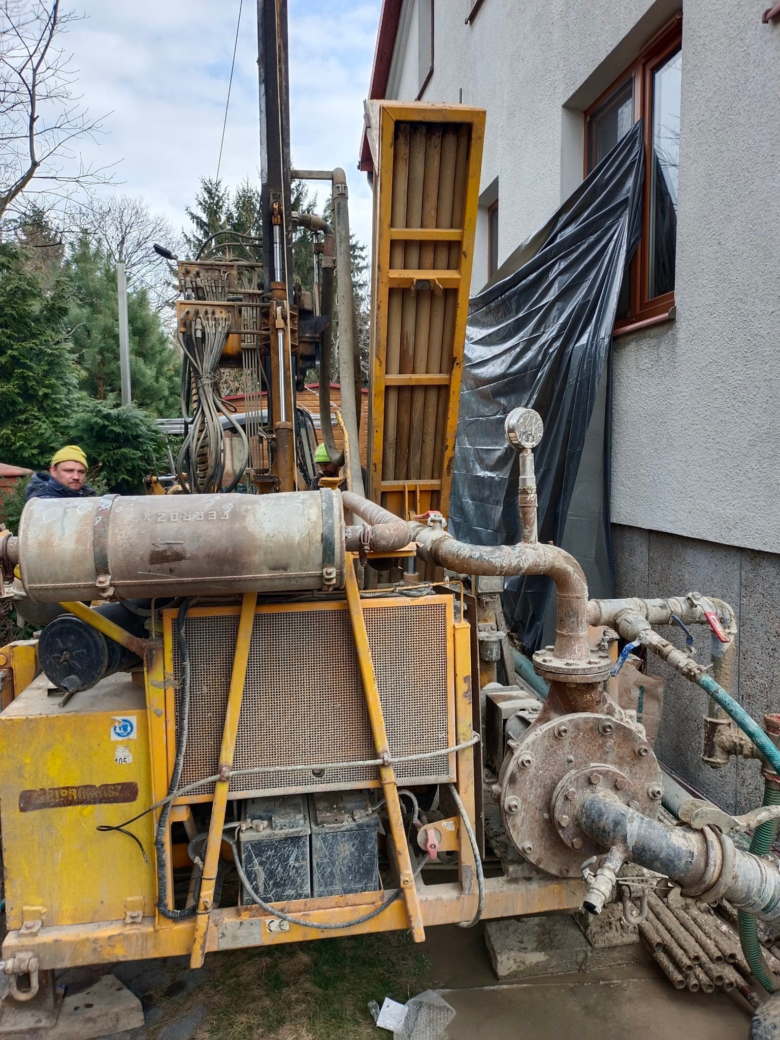 Żółta wiertnica z operatorem w żółtym kasku podczas pracy przy ścianie budynku, okno zabezpieczone czarną folią, widoczne przewody i rury.