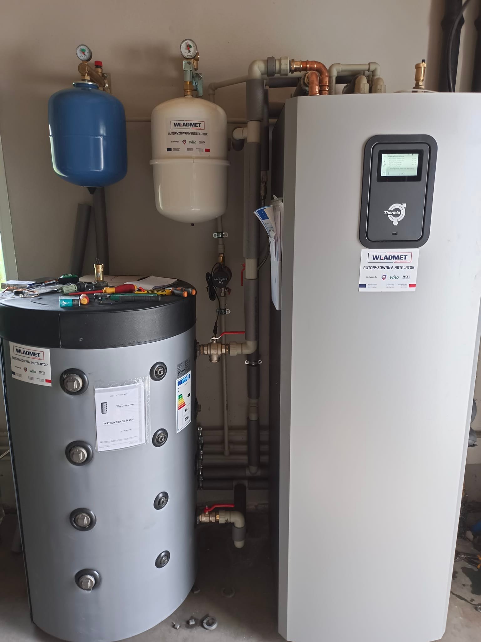 Zestaw urządzeń grzewczych z widocznymi zbiornikami wyrównawczymi, zasobnikiem ciepłej wody i jednostką sterującą Thermia, z narzędziami leżącymi na górnej powierzchni zasobnika.