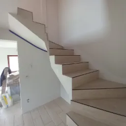 Klatka schodowa w trakcie wykańczania w Łętowni: drewnopodobne płytki na schodach z czarnymi krawędziami, ściany przygotowane do malowania, widoczne zabezpieczenia taśmą malarską.