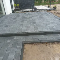 Szara kostka brukowa ułożona na tarasie z dwoma stopniami, widok z góry, obrzeża z ciemnej kostki, w tle dom i ogrodzenie z siatki.