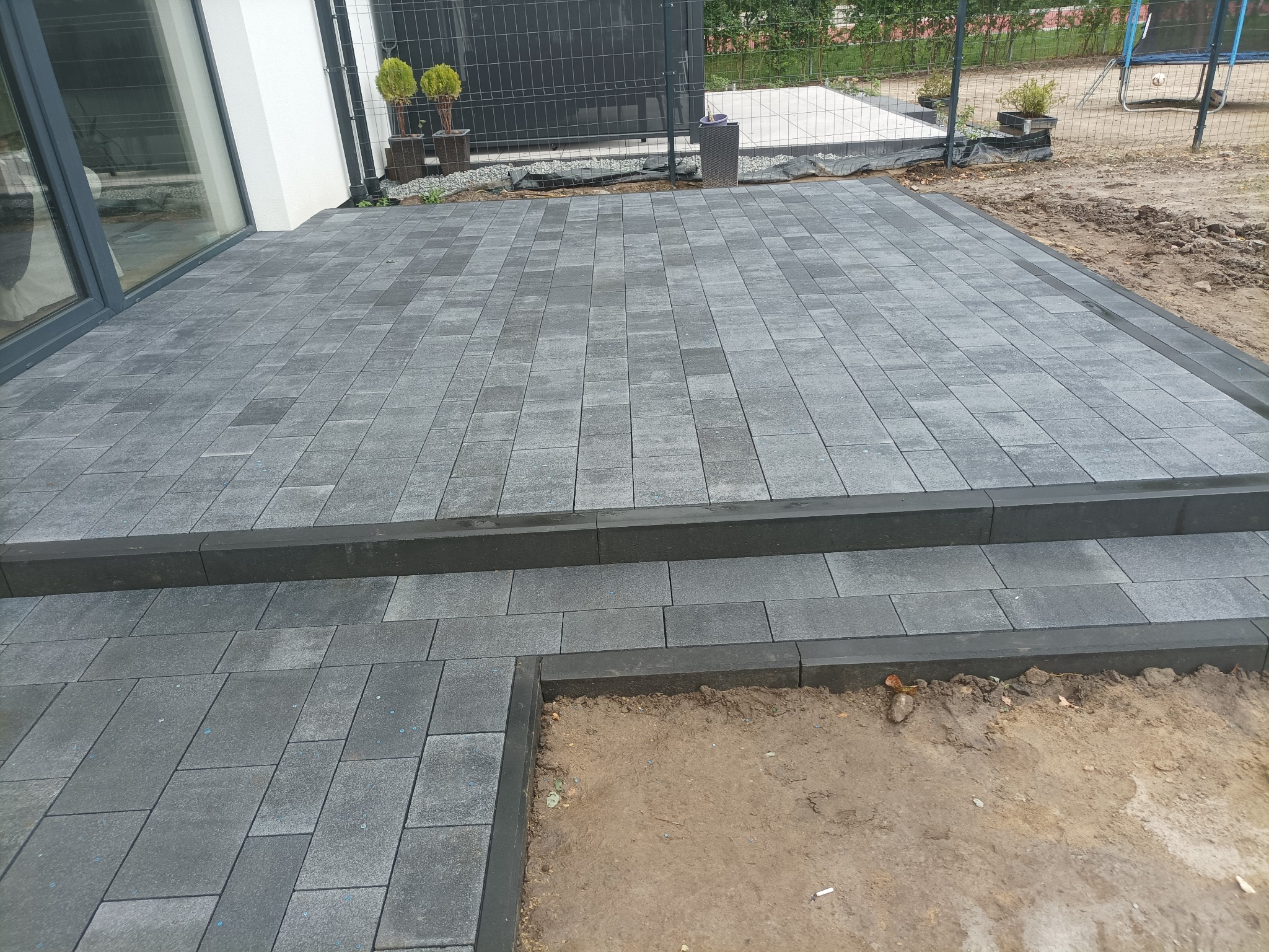 Szara kostka brukowa ułożona na tarasie z dwoma stopniami, widok z góry, obrzeża z ciemnej kostki, w tle dom i ogrodzenie z siatki.