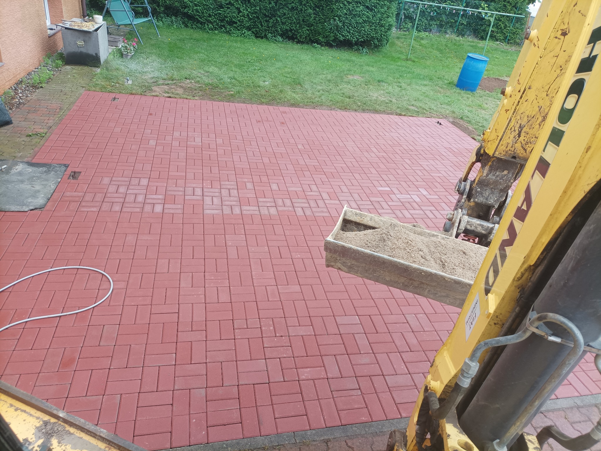 Czerwona kostka brukowa na posesji, z boku widoczny fragment koparki z piaskiem.