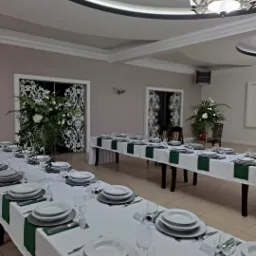 Uroczysta sala bankietowa z dwoma długimi stołami udekorowanymi białymi obrusami, zielonymi bieżnikami, elegancką zastawą i wysokimi kompozycjami kwiatowymi w wazonach, widoczne ozdobne drzwi...
