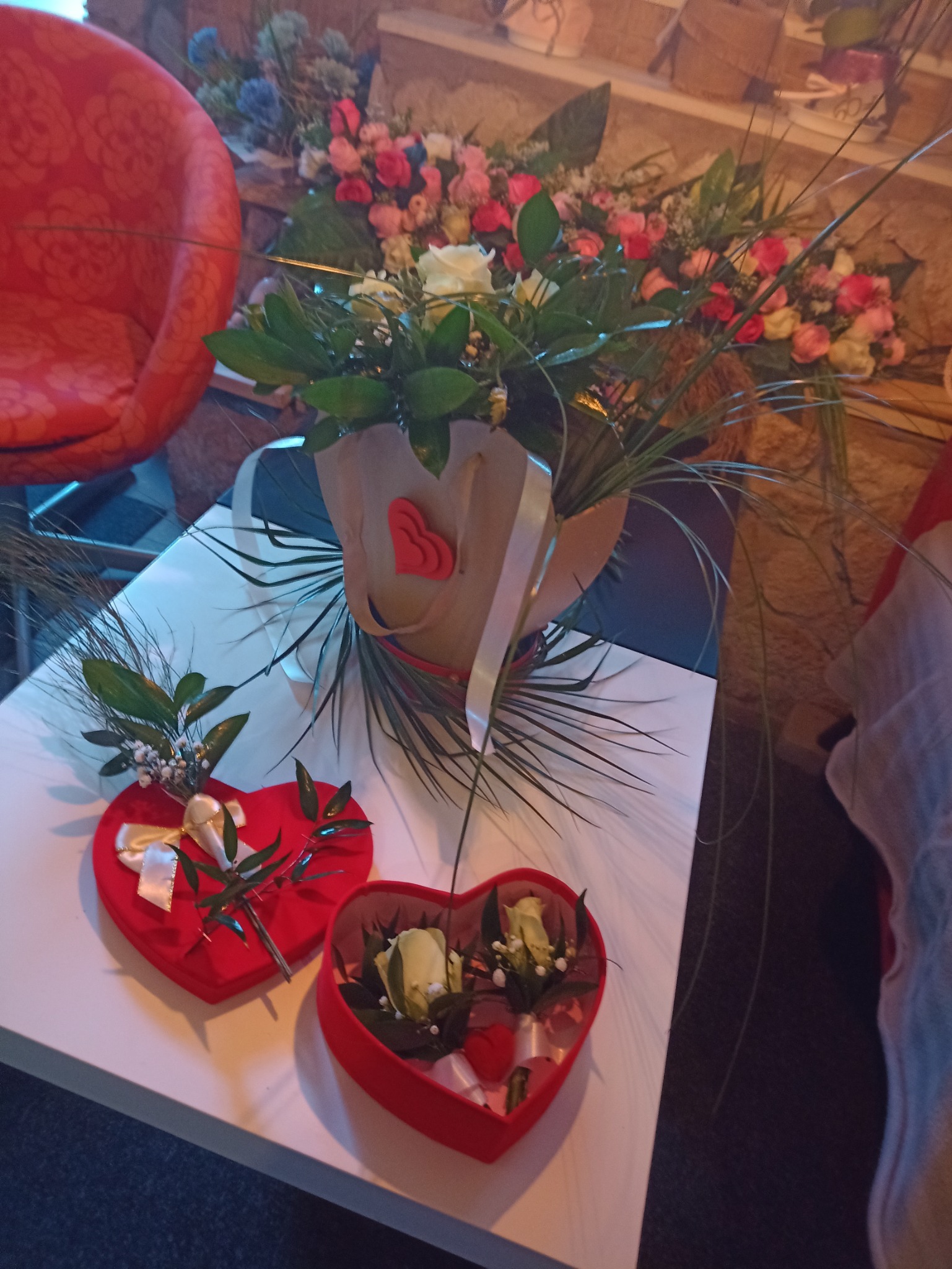 Kompozycja kwiatowa w brązowym wazonie z sercem, bukiet z różowych i białych kwiatów oraz dwa czerwone pudełka w kształcie serca z białymi różami i dekoracjami na białym stole.