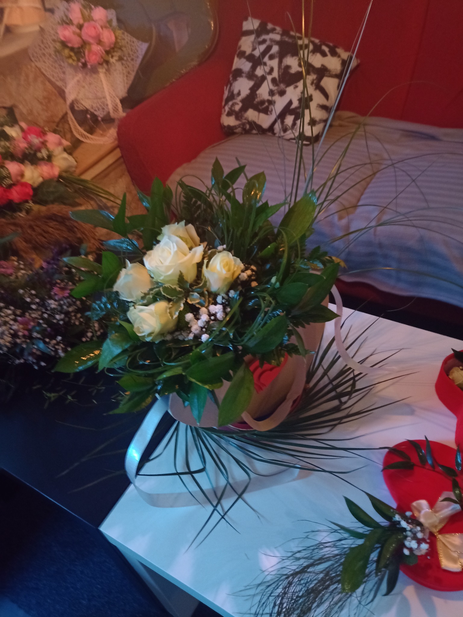 Bukiet kremowych róż z zielonymi liśćmi i drobnymi białymi kwiatami w ozdobnym opakowaniu, ustawiony na stole obok innych bukietów i dekoracji.