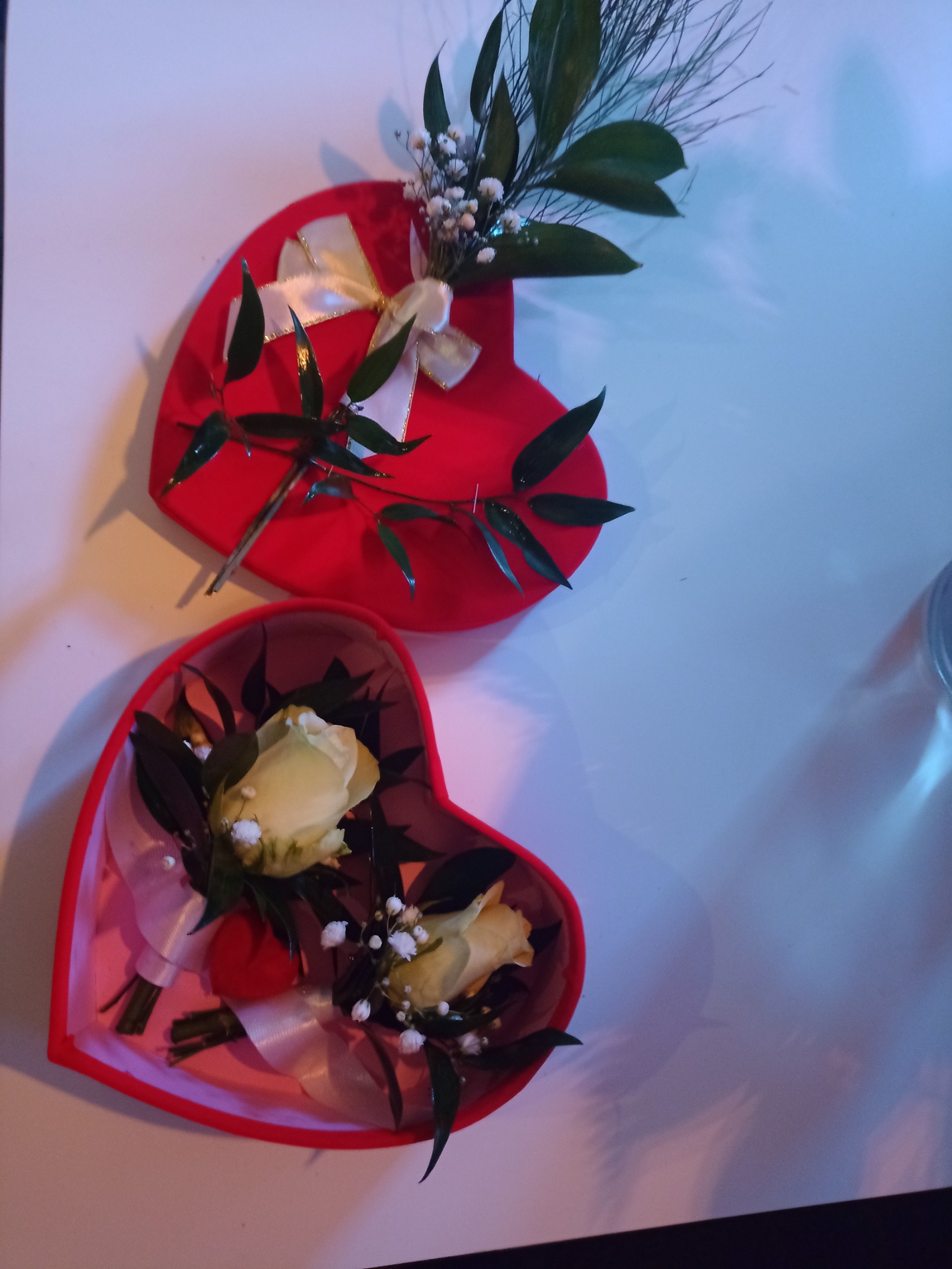 Dwa czerwone pudełka w kształcie serca, jedno zamknięte z białą kokardą i zielonymi gałązkami, drugie otwarte z dwiema białymi różami, wstążką i drobnymi białymi kwiatkami w środku.