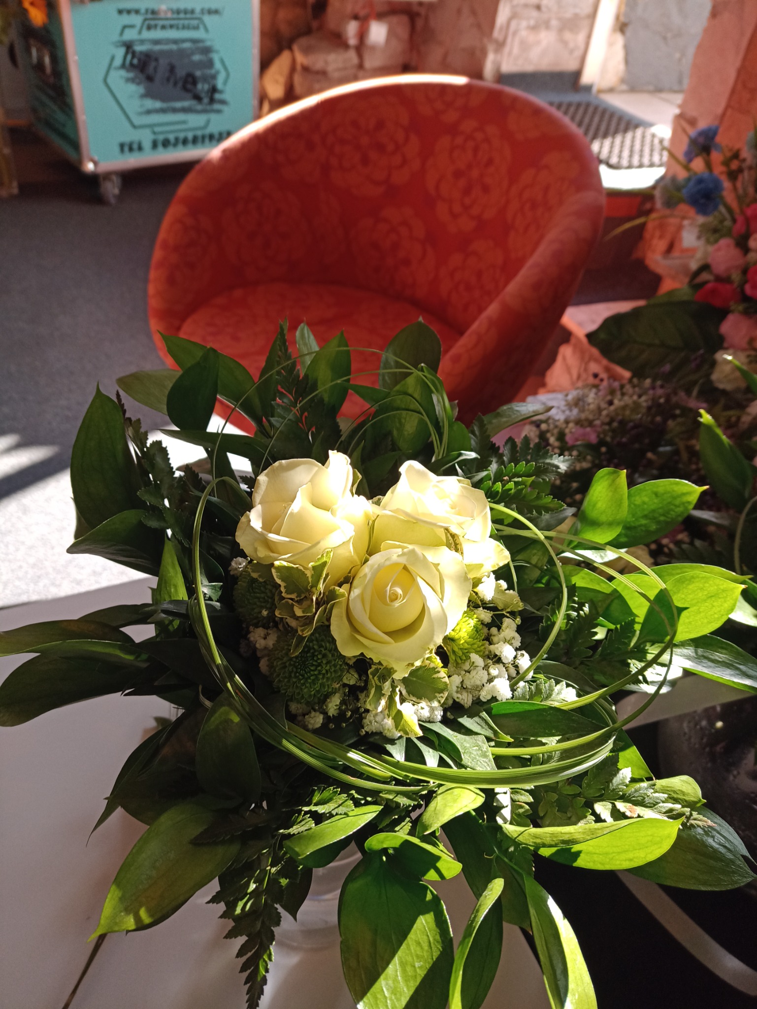 Białe róże w bukiecie z zielonymi dodatkami na stole, w tle pomarańczowy fotel i fragment wózka z reklamą.