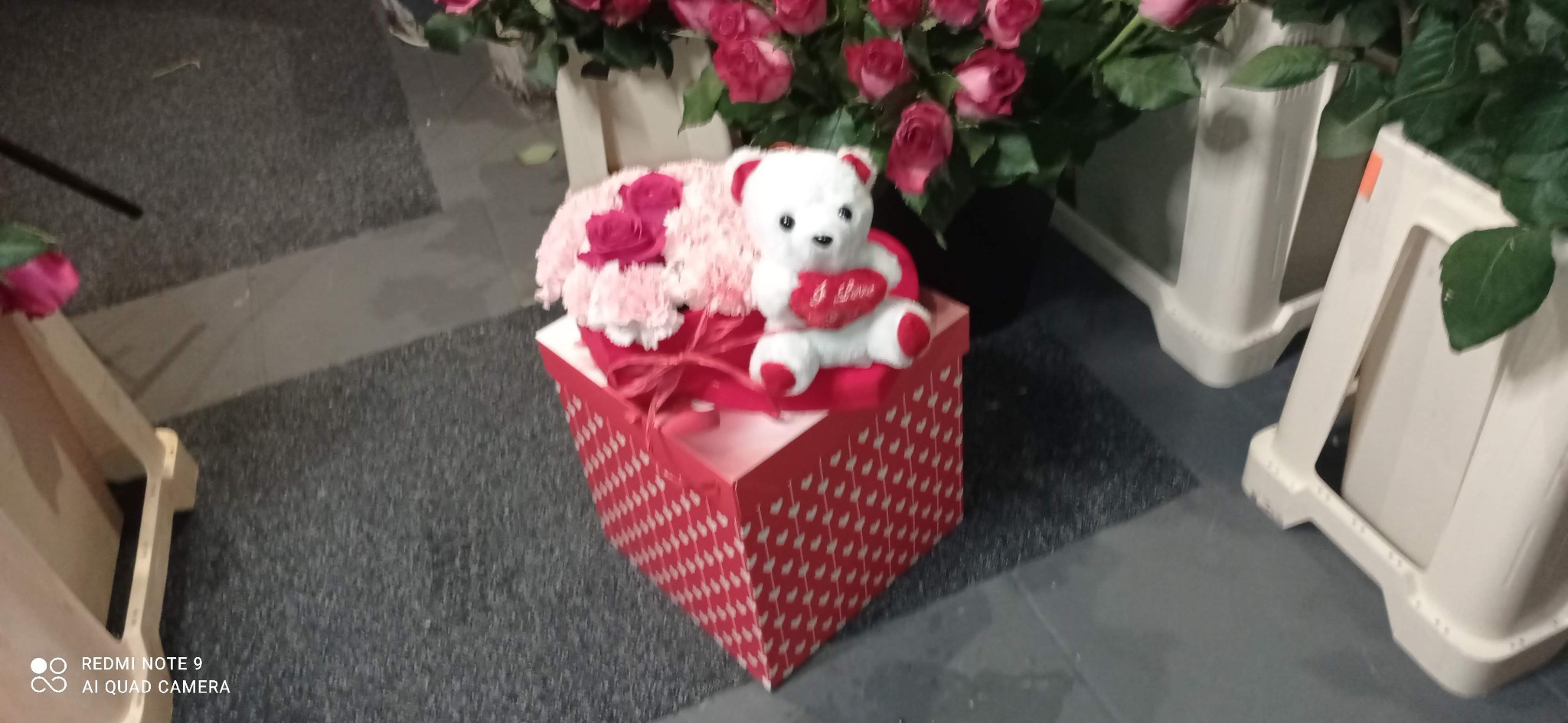 Kompozycja kwiatowa w czerwonym pudełku z serduszkami, ozdobiona pluszowym misiem trzymającym serce z napisem 'I love you', otoczona różami i innymi kwiatami.