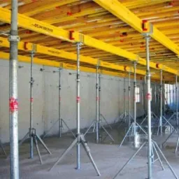 Wnętrze budynku w trakcie budowy, widoczne metalowe podpory podtrzymujące żółte, drewniane szalunki stropowe.