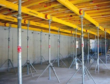 Wnętrze budynku w trakcie budowy, widoczne metalowe podpory podtrzymujące żółte, drewniane szalunki stropowe.