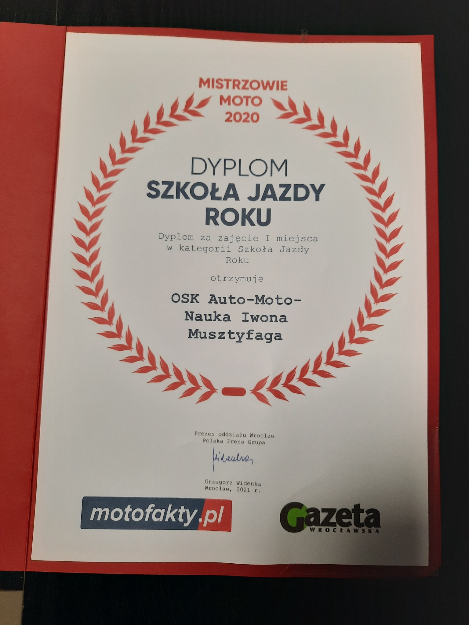 Dyplom dla OSK Auto-Moto-Nauka Iwona Musztyfaga za zajęcie I miejsca w kategorii Szkoła Jazdy Roku, otoczony czerwoną ramką i wieńcem z liści, z logo motofakty.pl i Gazety Wrocławskiej w dolnej...