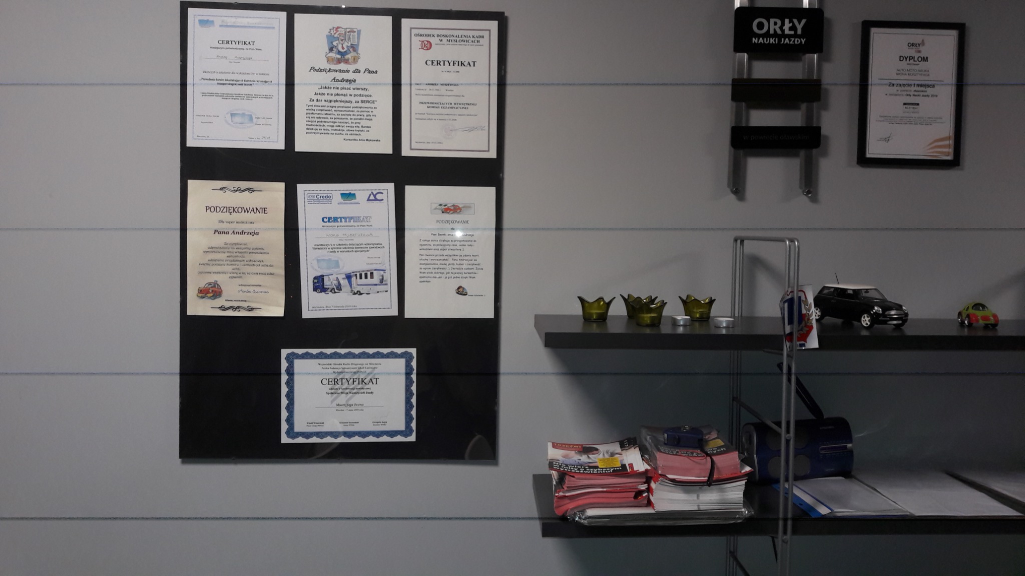 Ściana z certyfikatami i podziękowaniami dla instruktora, półki z modelami samochodów i materiałami biurowymi, widoczne logo firmy nauki jazdy.