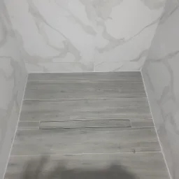 Nowoczesna kabina prysznicowa z szarymi płytkami imitującymi drewno na podłodze i białymi płytkami imitującymi marmur na ścianach, z odpływem liniowym.