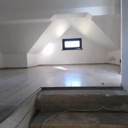 Wykończone poddasze z drewnianą podłogą, oknem dachowym i widoczną drewnianą belką konstrukcyjną, schody betonowe prowadzą na poddasze, widoczne kable elektryczne.