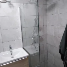 Nowoczesna łazienka z kabiną prysznicową typu walk-in, umywalką nablatową na szafce z drewnopodobnym frontem i szarą, kwadratową glazurą ścienną.