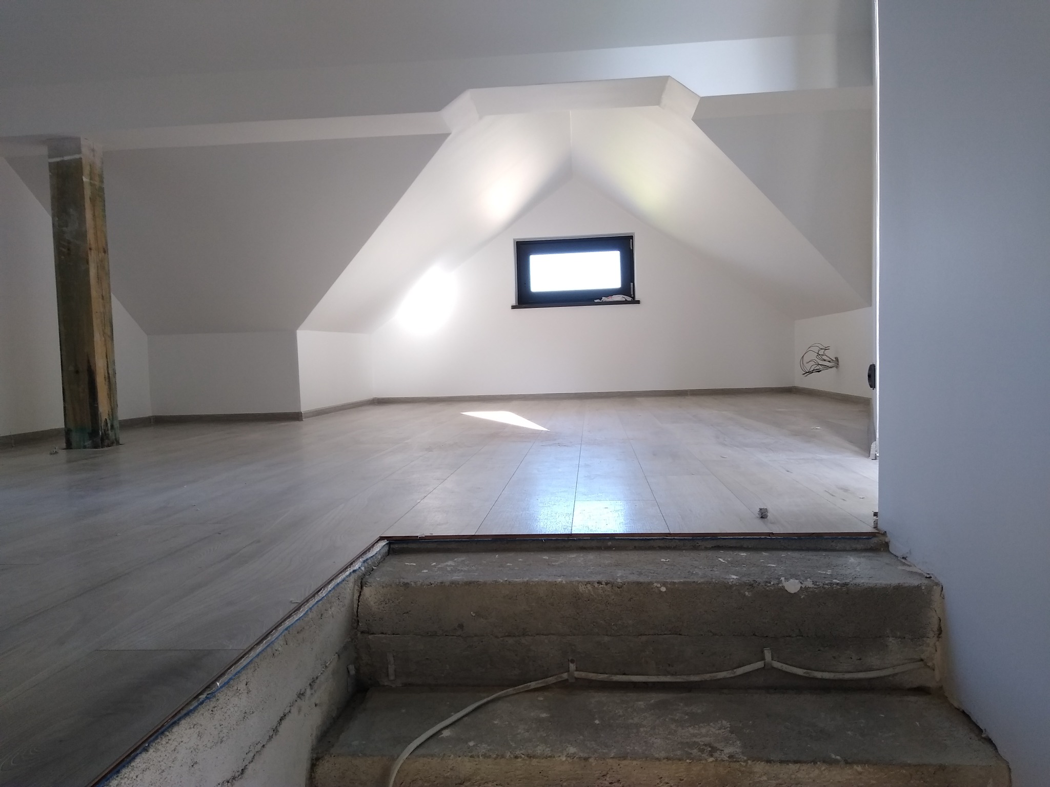 Wykończone poddasze z drewnianą podłogą, oknem dachowym i widoczną drewnianą belką konstrukcyjną, schody betonowe prowadzą na poddasze, widoczne kable elektryczne.