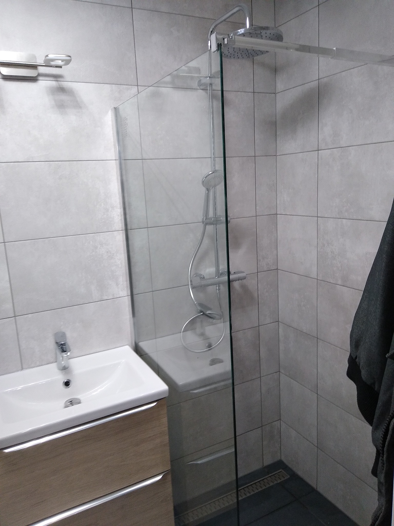 Nowoczesna łazienka z kabiną prysznicową typu walk-in, umywalką nablatową na szafce z drewnopodobnym frontem i szarą, kwadratową glazurą ścienną.