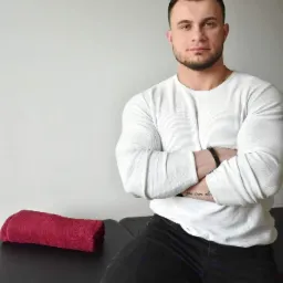 Mężczyzna w białej bluzce z długim rękawem, z założonymi rękami, siedzący na czarnym stole obok czerwonego ręcznika.