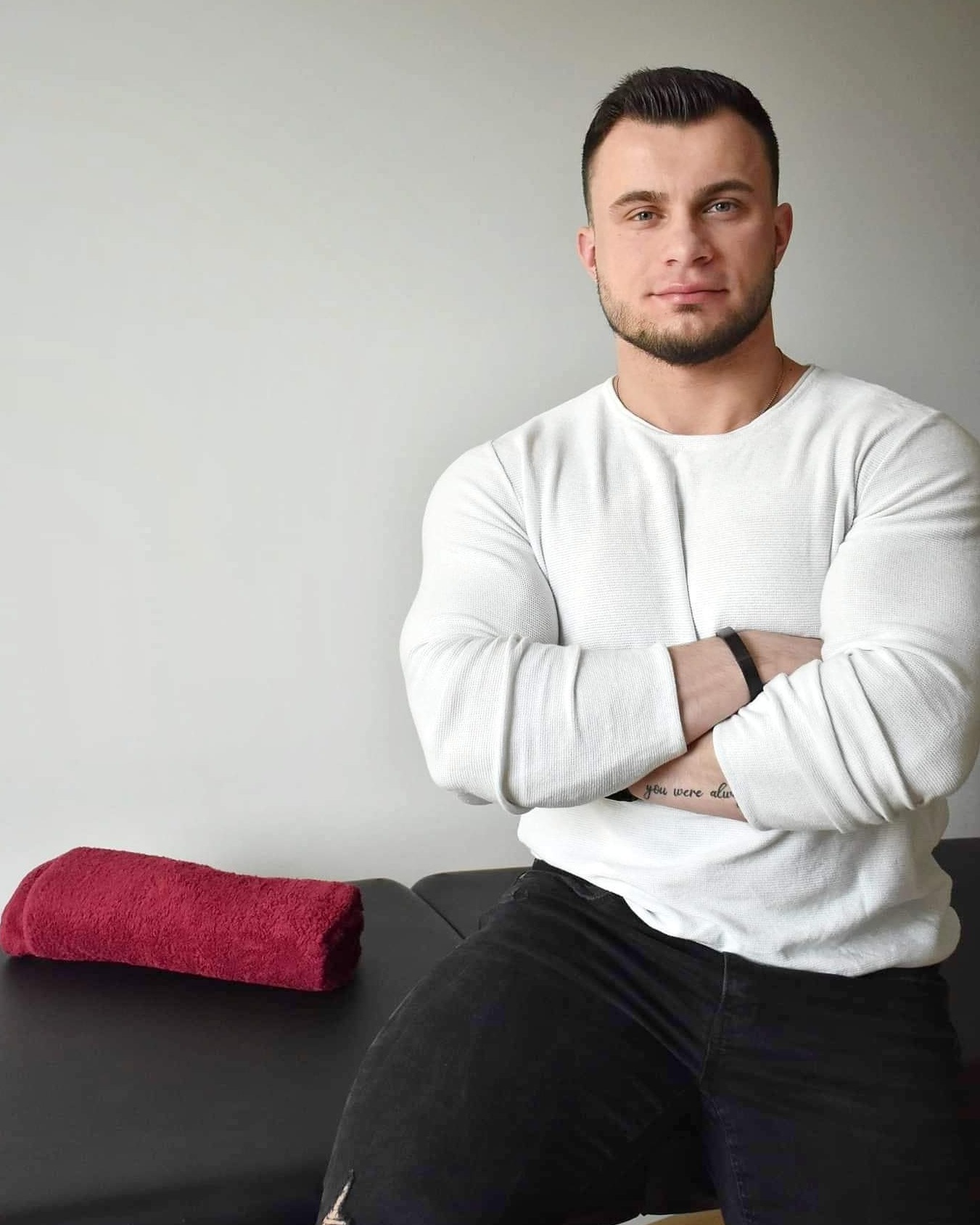 Mężczyzna w białej bluzce z długim rękawem, z założonymi rękami, siedzący na czarnym stole obok czerwonego ręcznika.