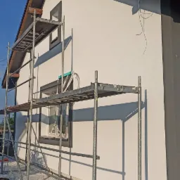 Świeżo otynkowana ściana domu w Tucholi, widoczna z perspektywy rusztowania, z zabezpieczoną folią podmurówką i wiaderkiem z tynkiem silikonowym w pobliżu.