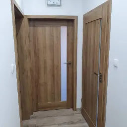 Dwa drewniane drzwi wewnętrzne z przeszkleniami, zamontowane w białym korytarzu, z widocznym zegarem na ścianie i panelami podłogowymi imitującymi drewno.