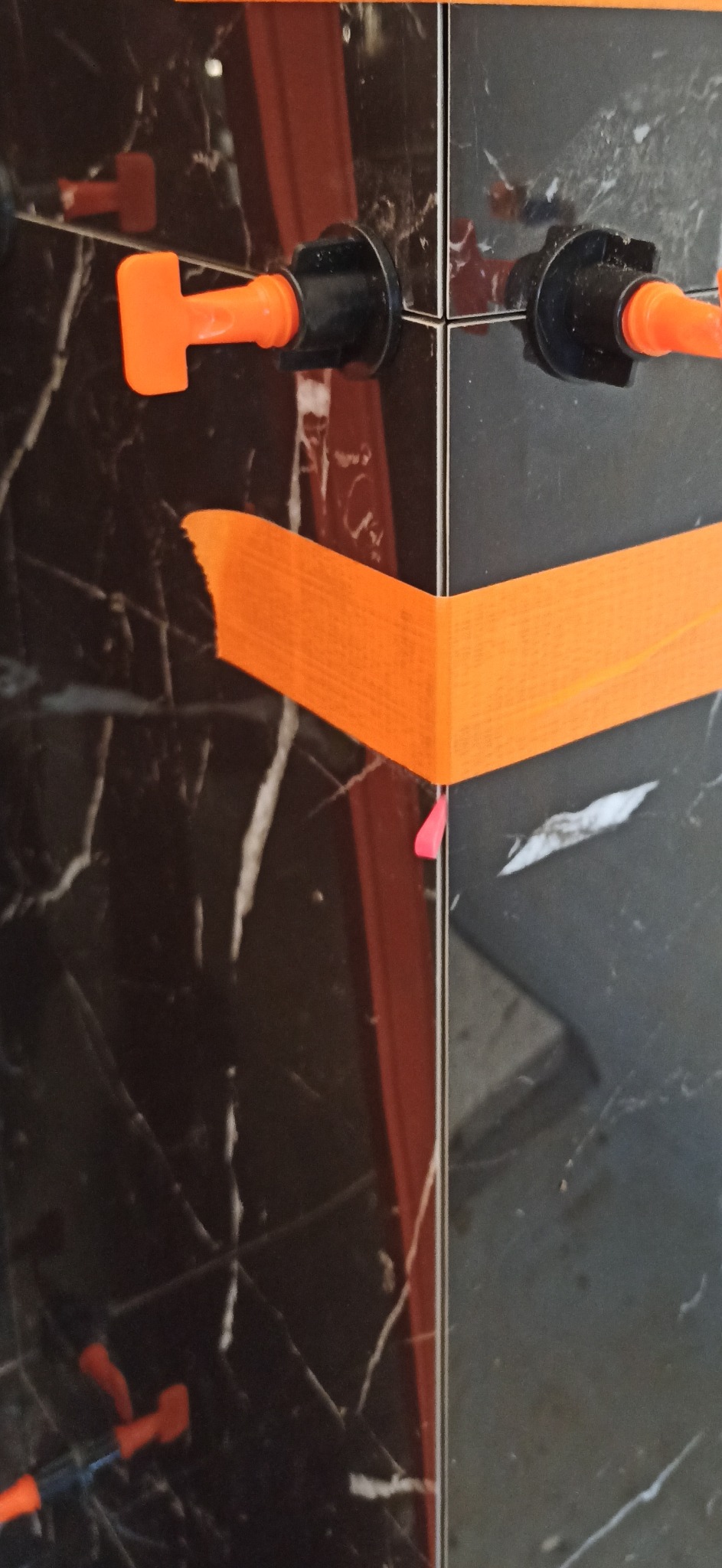 Układanie płytek imitujących marmur w narożniku, z użyciem pomarańczowej taśmy maskującej i systemu poziomowania płytek z pomarańczowymi klinami.