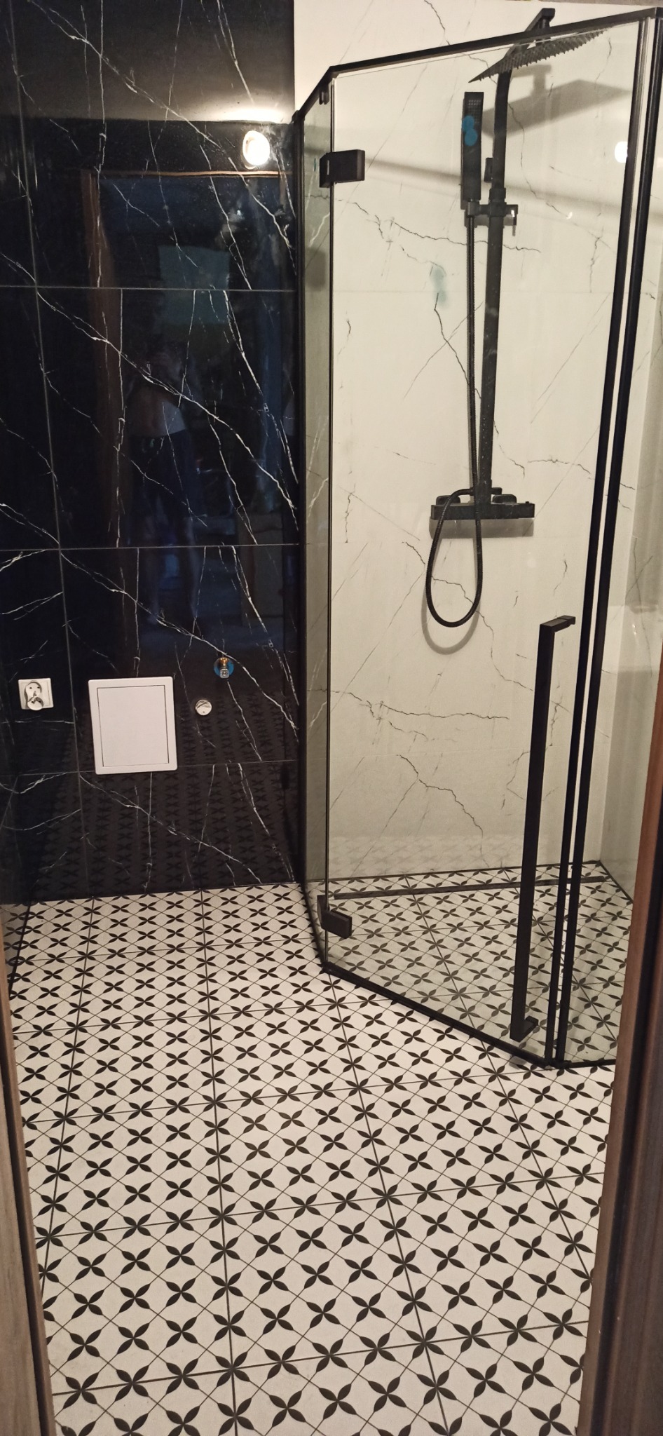 Nowoczesna kabina prysznicowa z czarną armaturą, marmurowe płytki na ścianach i podłodze, geometryczny wzór na podłodze, czarne płytki imitujące marmur na ścianie z ukrytym stelażem WC.