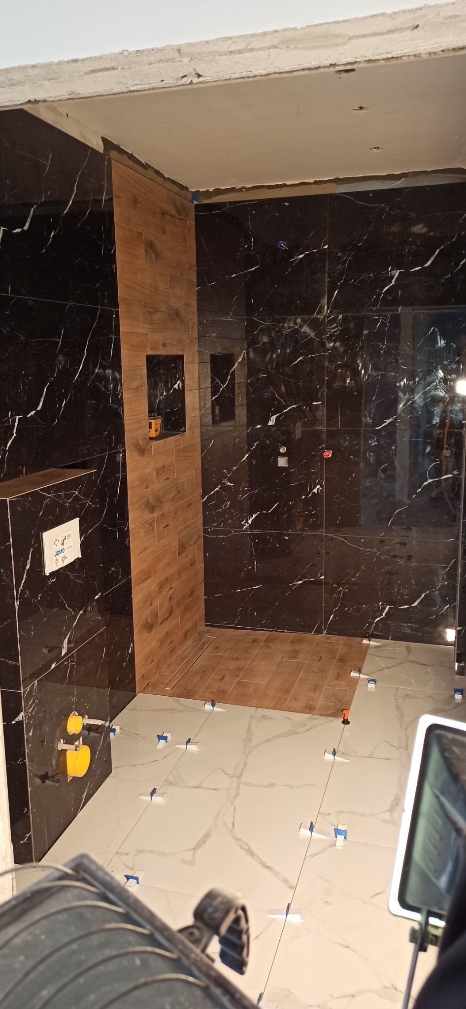 Łazienka w trakcie remontu: czarne płytki imitujące marmur na ścianach, drewnopodobne płytki w strefie prysznica, jasne płytki podłogowe z krzyżykami dystansowymi, widoczne elementy instalacji...