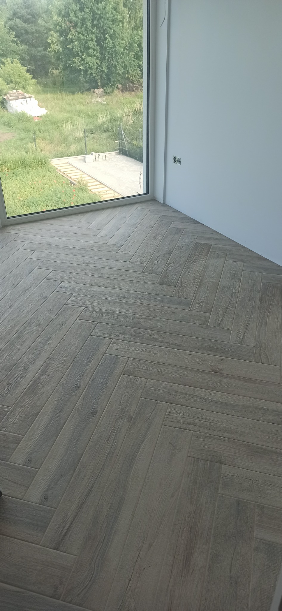 Ułożona w jodełkę podłoga z płytek imitujących drewno w nowym wnętrzu, widok na zewnątrz przez duże okno.