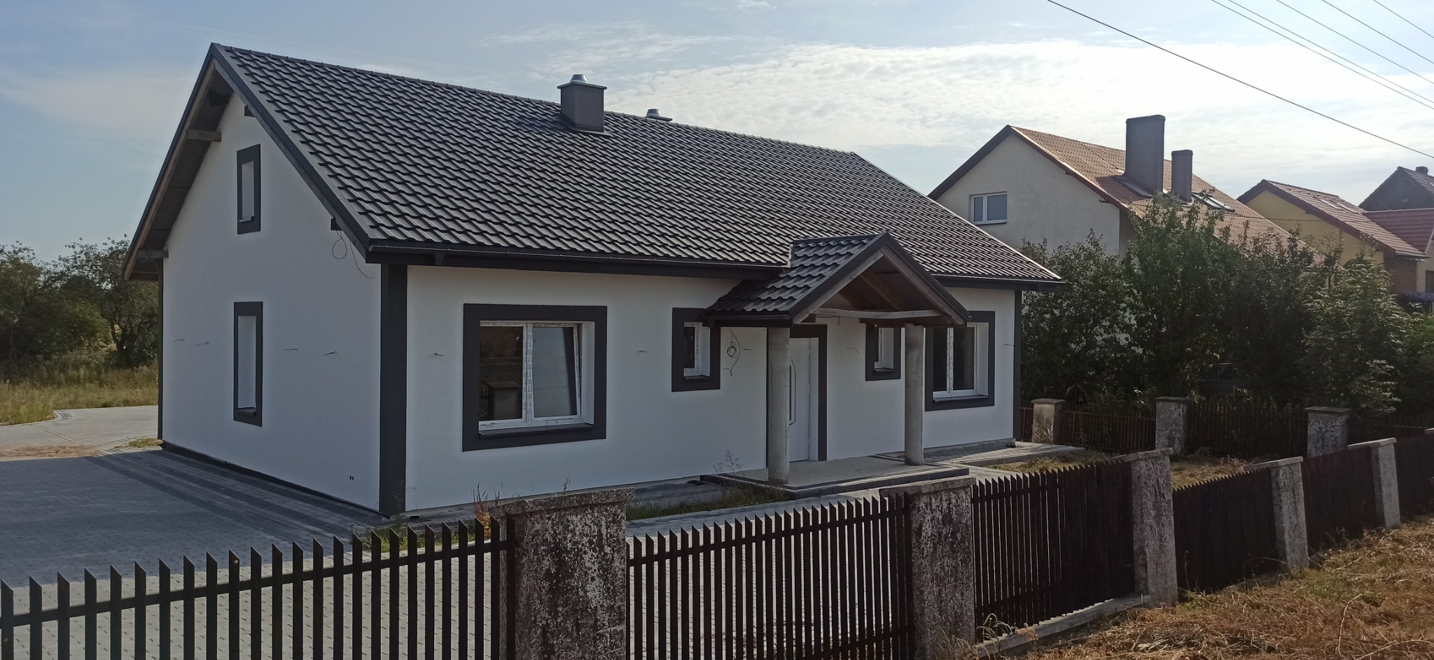 Nowo wybudowany dom jednorodzinny z białymi ścianami i ciemną dachówką, widoczny zza ciemnego, drewnianego płotu z betonowymi słupkami.