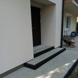 Grupa ZieLiński - Wykończone schody wejściowe z szarej kostki z czarnym obramowaniem, prowadzące do ciemnych drzwi wejściowych. W tle widoczna taczka z betoniarką.