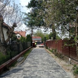 Grupa ZieLiński - Długa, wybrukowana droga dojazdowa wykonana z szaro-czarnej kostki, otoczona ogrodzeniami z drewna i metalu, z widoczną ciężarówką w oddali i pracownikiem w kamizelce odblaskowej.