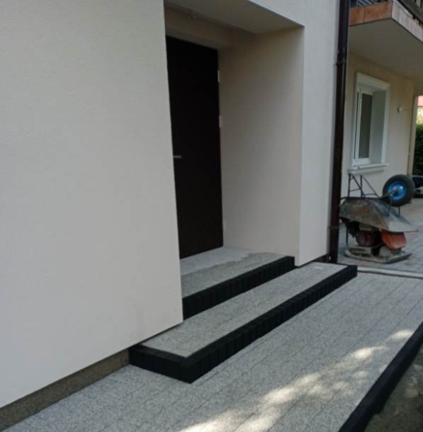 Wykończone schody wejściowe z szarej kostki z czarnym obramowaniem, prowadzące do ciemnych drzwi wejściowych. W tle widoczna taczka z betoniarką.